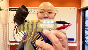 ごんた屋代表の徳久（のりひさ）が、自社開発の「バックフラッシャー動作確認ボード」を手に、製品の確実な動作と品質を解説している様子。