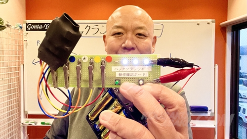 ごんた屋代表の徳久（のりひさ）が、自社開発の「バックフラッシャー動作確認ボード」を手に、製品の確実な動作と品質を解説している様子。